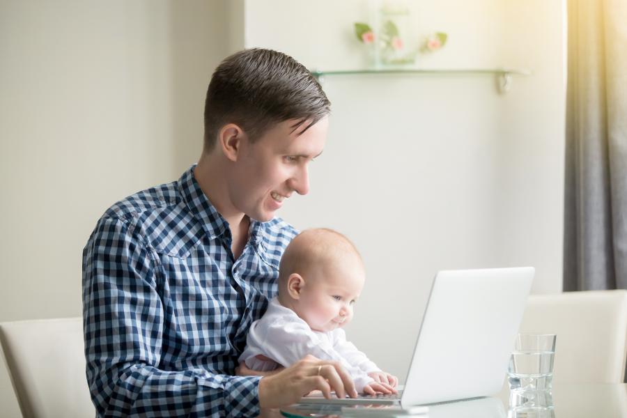 Man in shirt sat at desk using a laptop, with baby sat on his lap